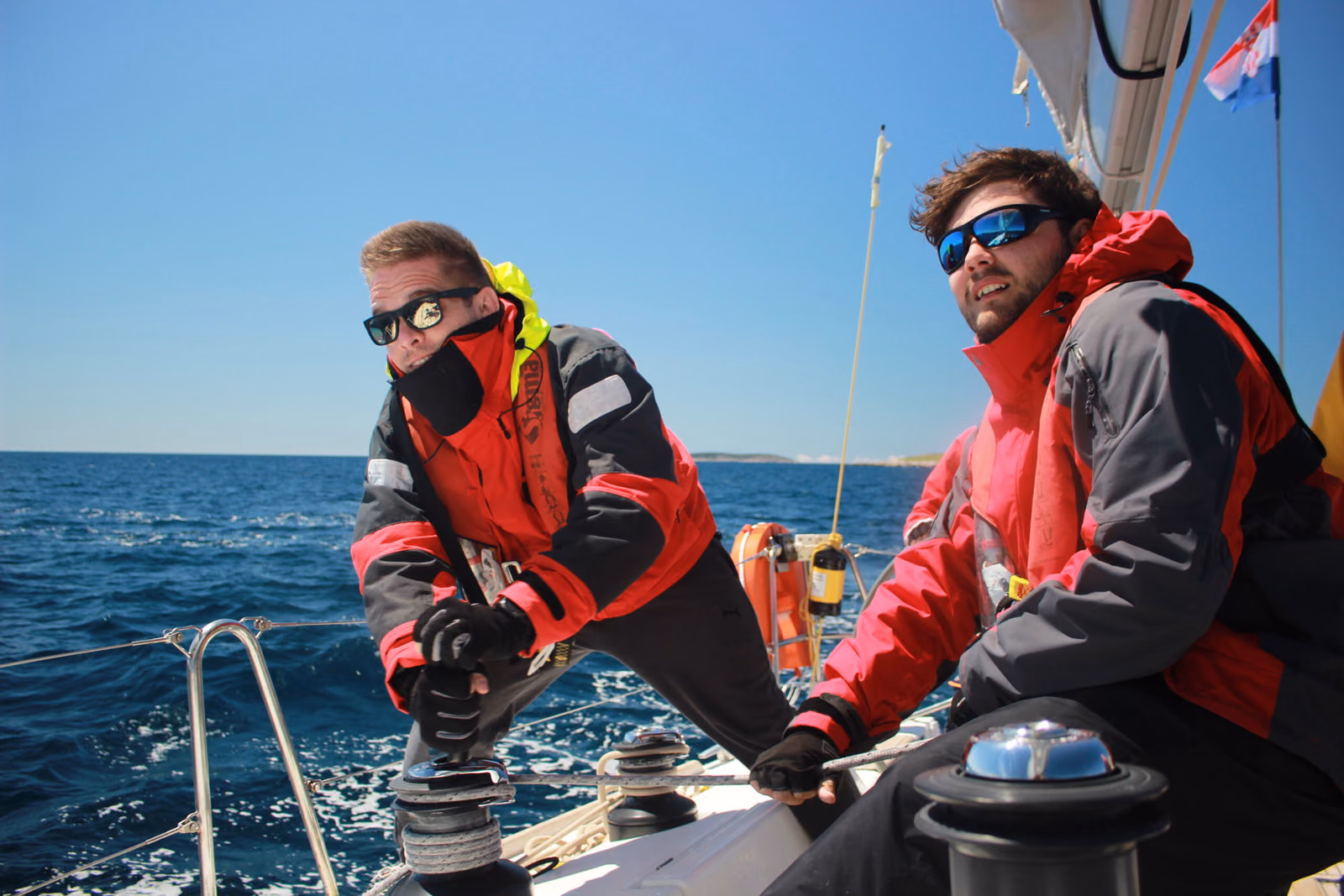 Two students in sailing jackets working the winches on a yacht during an RYA course